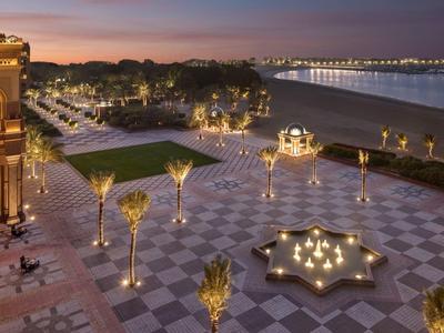 Elegante Terrasse mit Palmen und beleuchtetem Brunnen am Wasser bei Sonnenuntergang.