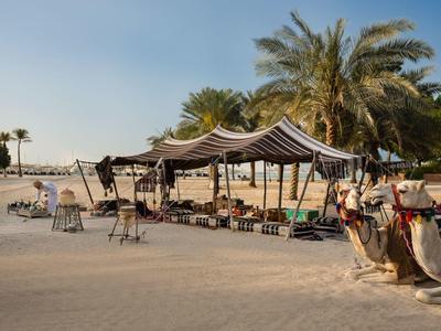 Wüstenzelt mit Kamel und Stühlen auf sandigem Boden unter blauem Himmel.