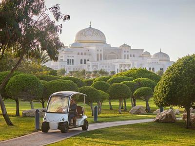 Golfwagen fährt auf einem Weg durch gepflegten Park mit weißen Palastgebäuden im Hintergrund.
