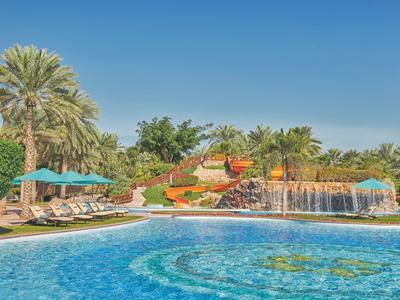 Großer Pool mit Wasserrutsche, Wasserfall und Liegestühlen in tropischer Gartenanlage.