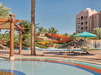 Hotelpool mit Liegestühlen, Grillplatz, Palmen und Wasserrutschen unter blauem Himmel.