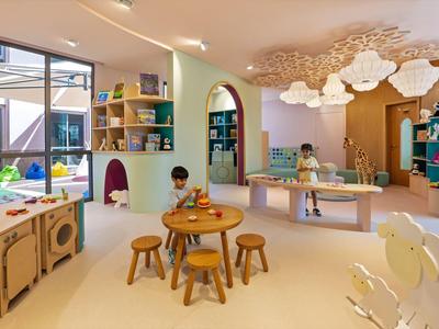 Heller Kinderraum mit Tischen, Stühlen, großen Fenstern und spielenden Kindern.