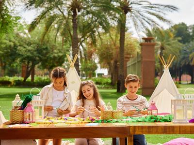 Drei Kinder sitzen im Freien an einem Holztisch mit Bastelmaterial und tipis im Hintergrund.