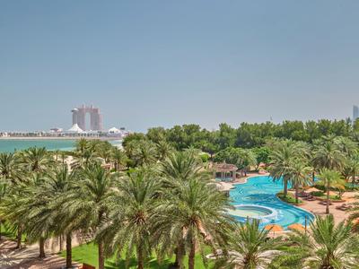 Blick auf einen tropischen Hotelpool mit Palmen und Meer im Hintergrund bei klarem Himmel.