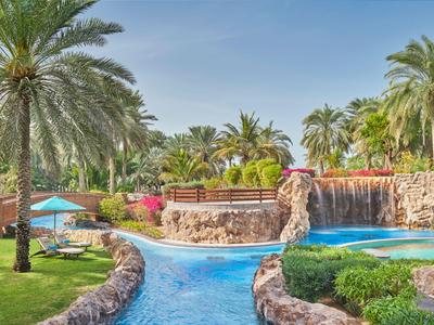 Außenpool mit Wasserfall, blauer Liegestuhl und Palmen in einer grünen Gartenanlage.