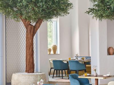 Modernes Café mit blauen und cremefarbenen Stühlen um Holz- und Marmor-Tische, dekoriert mit großen Pflanzen.