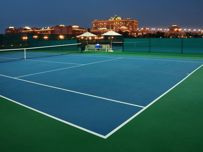 Beleuchteter Tennisplatz mit Hotelgebäuden im Hintergrund bei Nacht.