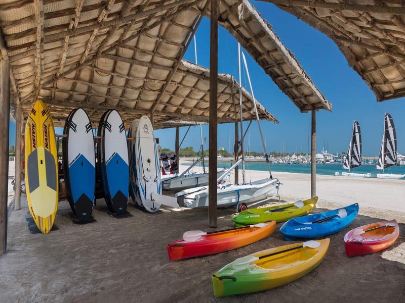Bunte Kajaks und Surfboards unter einem Strohdach am Strand bei sonnigem Himmel.