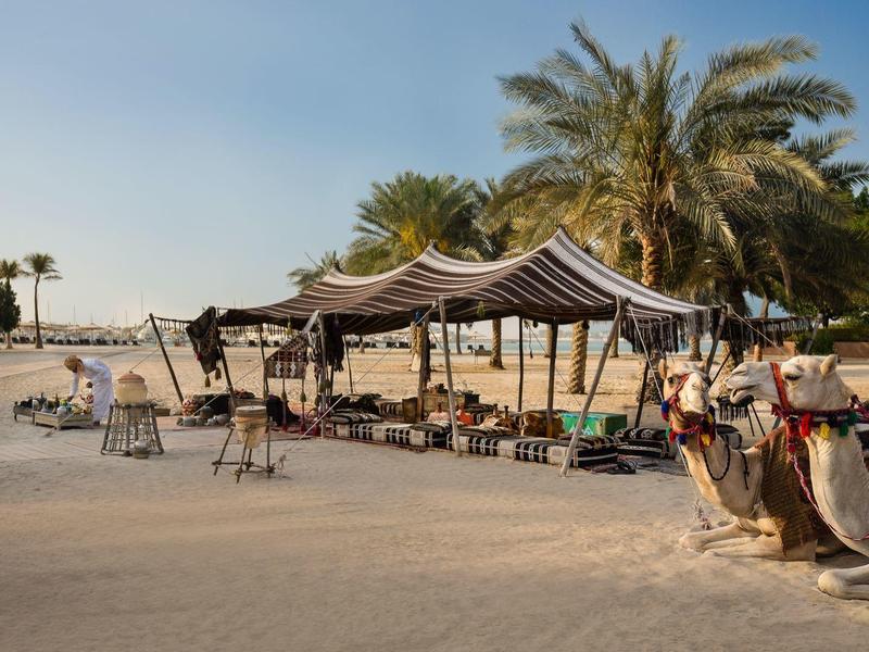 Wüstenzelt mit Kamel und Stühlen auf sandigem Boden unter blauem Himmel.