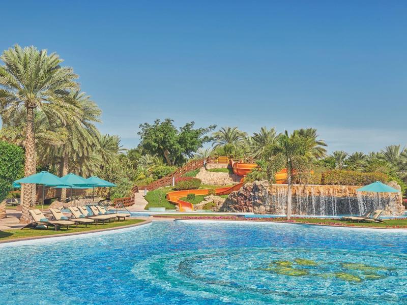Großer Pool mit Wasserrutsche, Wasserfall und Liegestühlen in tropischer Gartenanlage.