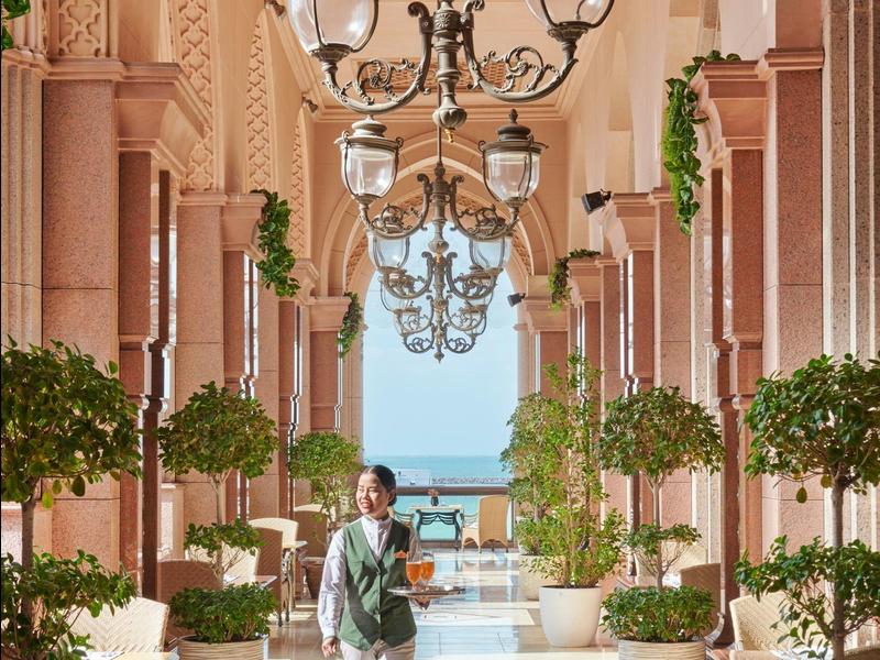 Ein eleganter Gang mit Kronleuchtern und großen Pflanzen entlang der Wände und Blick aufs Meer.
