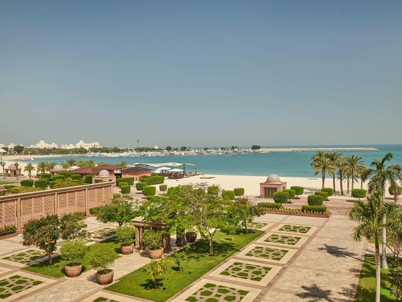Blick auf eine gepflegte Gartenanlage mit Palmen und Strand am Meer unter klarem Himmel.