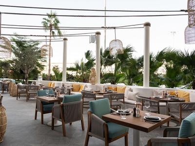 Außenrestaurant mit Holztischen, gepolsterten Stühlen und Palmen im Hintergrund.