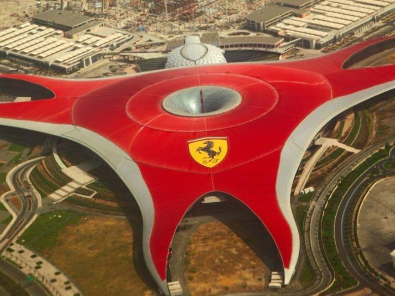 Großes, rotes, futuristisches Gebäude mit Ferrari-Logo, umgeben von Straßen und grünen Flächen.