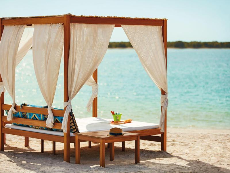 Holz-Pavillon mit weißen Vorhängen und Kissen am Sandstrand neben blauem Meer.