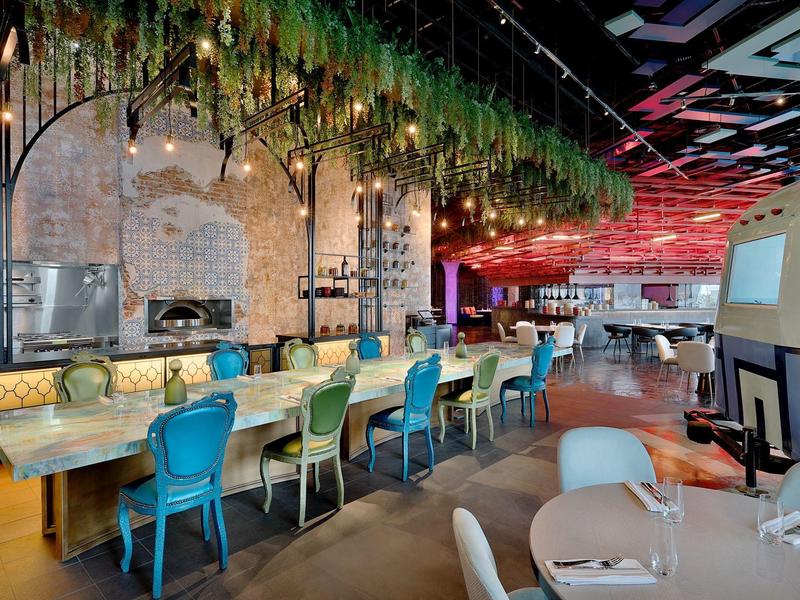 Moderner Gastraum mit bunten Stühlen, grünen Hängepflanzen und beleuchtetem Holztresen.