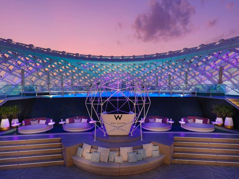 Moderner Außenbereich mit bunter LED-Beleuchtung, Lounge-Möbeln und zentraler runder Bühne bei Abenddämmerung.