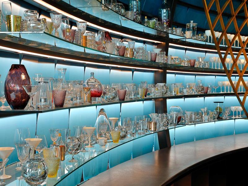 Vitrine mit geschwungenen Glasregalen, gefüllt mit vielfältigen, bunten Gläsern und Karaffen.