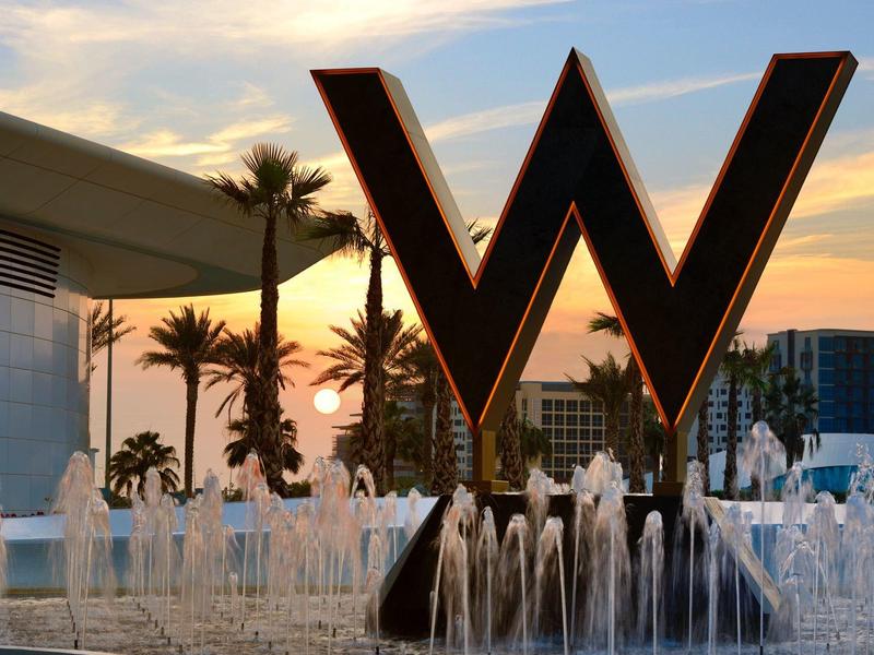 Großes W-Logo am Springbrunnen vor Hotelgebäude mit Palmen im Sonnenuntergang.
