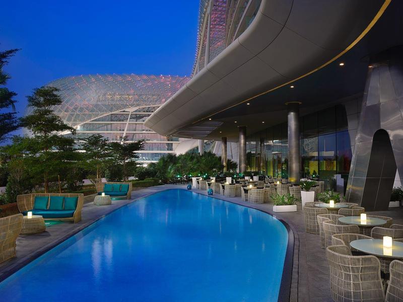 Moderner Außenpool mit blauem Wasser, umgeben von Tischen, Stühlen und begrünter Terrasse bei Nacht.