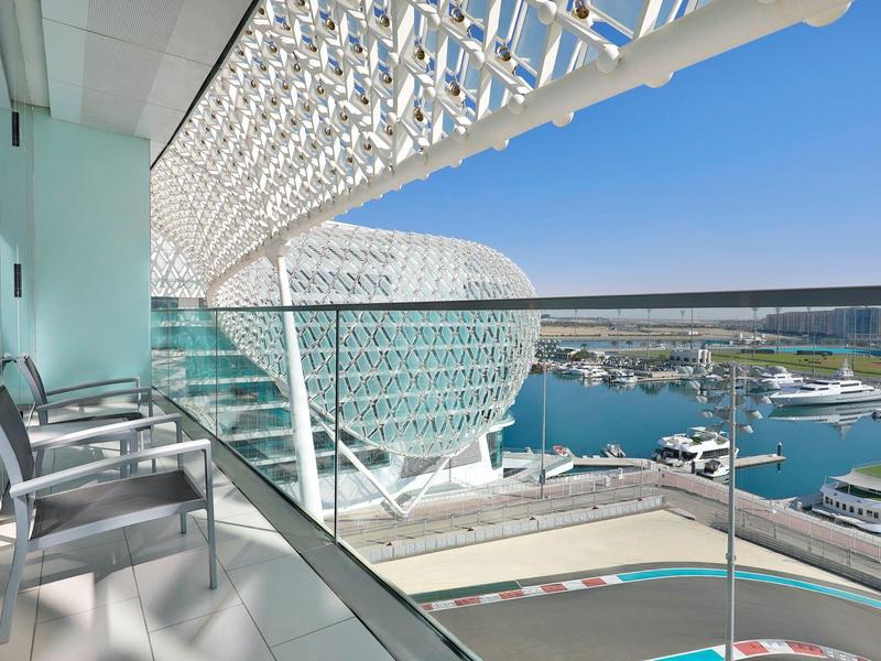 Balkon mit Stühlen und Glasgeländer, Blick auf moderne runde Architektur und Hafen bei klarem Himmel.