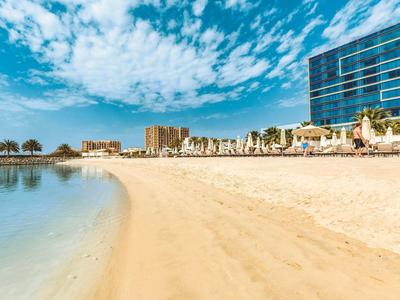 Strand mit hellem Sand, ruhigem Wasser, blauen Himmel und modernen Gebäuden am Rand.