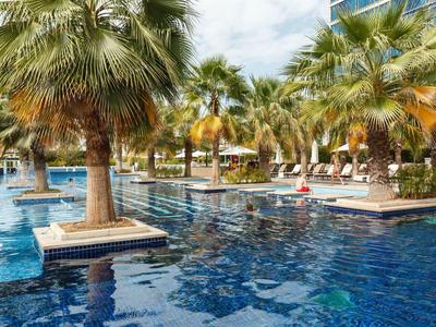 Großer Außenpool mit blauer Wasseroberfläche und Inseln mit Palmen, umgeben von Liegestühlen.