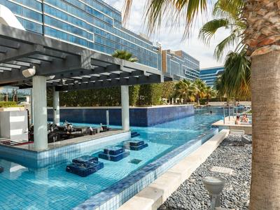 Moderner Außenpool mit Schwimmbarsitzen in blauem Wasser, Palmen und Glasfassade im Hintergrund.