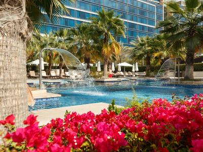 Ein Pool mit klarem Wasser, umgeben von Palmen und roten Blumen, vor einem modernen Glasgebäude.