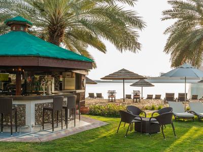 Outdoor bar with stools and seating under palm trees near sun loungers and umbrellas by the water.