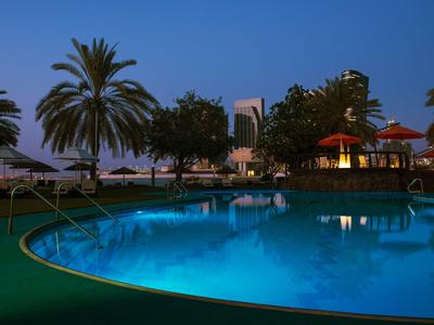 Abendaufnahme eines beleuchteten Pools mit Palmen und Hotelgebäuden im Hintergrund.