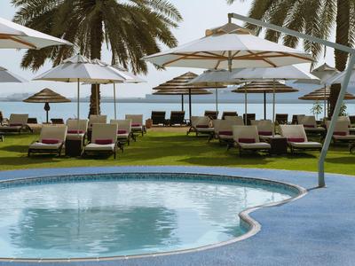 Runder Pool mit blauen Fliesen, Liegestühle und weiße Sonnenschirme, Palmen und Meer im Hintergrund.