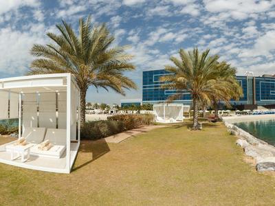 Freizeitbereich mit einer modernen, weißen Lounge-Box, Palmen und Wasser umgeben von Gebäuden.