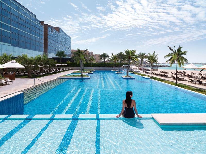 Großer blauer Pool mit Palmen und Liegestühlen, Person sitzt am Beckenrand bei sonnigem Himmel.
