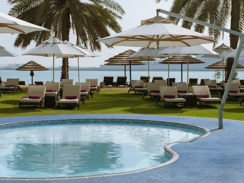 Runder Pool mit blauen Fliesen, Liegestühle und weiße Sonnenschirme, Palmen und Meer im Hintergrund.
