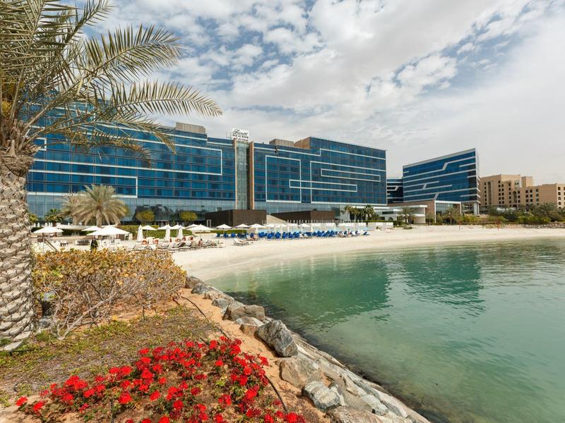 Moderner Gebäudekomplex mit Glasfassade an ruhigem Strand, Palmen und Blumen im Vordergrund.