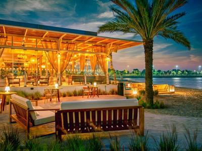 Strandbar bei Sonnenuntergang mit Holzmöbeln, Palmen und beleuchtetem Meer im Hintergrund.