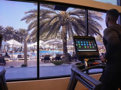 Blick aus dem Fitnessstudio auf Palmen und Poolbereich eines Resorts bei Sonnenuntergang.