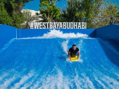 Person surft auf einer blauen künstlichen Welle vor grünem Hintergrund und blauem Himmel.