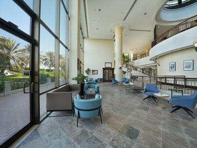 Großer heller Loungebereich mit hohen Fenstern, grauem Boden, blauen Sesseln und moderner Einrichtung.