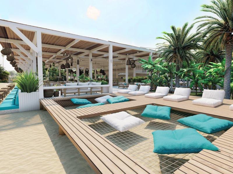 Moderner Strandclub mit Lounge-Bereich, Holzboden, blauen Kissen und Palmen im Hintergrund.