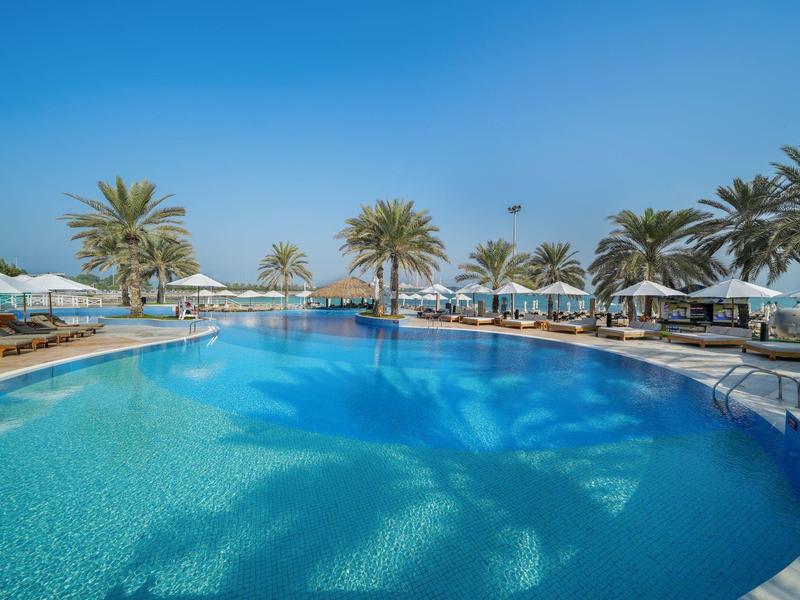 Großer, klarer Pool mit Palmen und weißen Sonnenschirmen unter blauem Himmel.