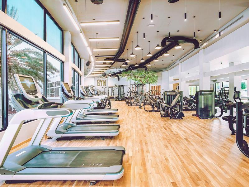 Großer Fitnessraum mit Laufbändern, Geräten und Holzboden, helle Fenster, hohe Decke.