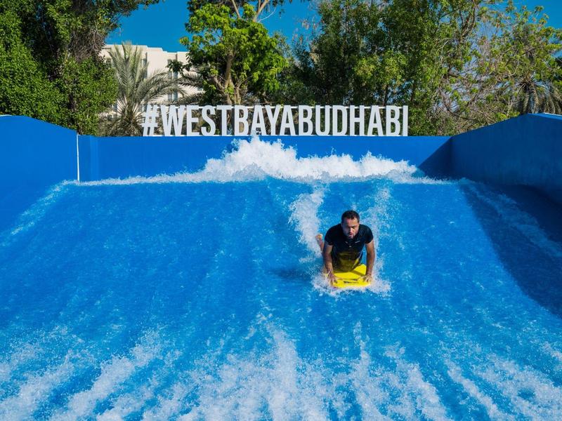 Person surft auf einer blauen künstlichen Welle vor grünem Hintergrund und blauem Himmel.