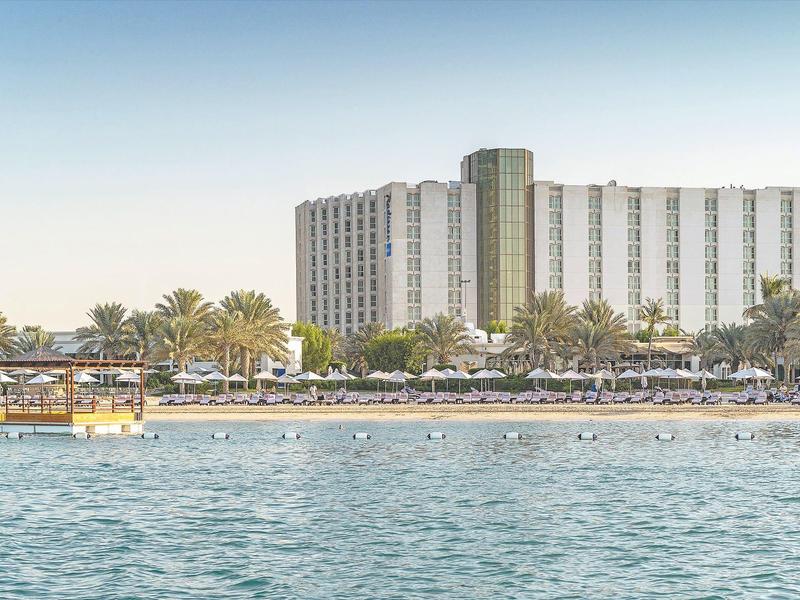 Hotelgebäude am Ufer mit Palmen und blauem Wasser im Vordergrund unter klarem Himmel.