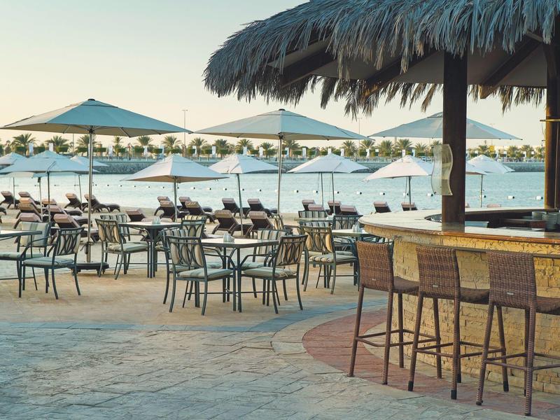 Strandbar und Tische mit Stühlen unter Sonnenschirmen am Sandstrand bei ruhigem Meer.