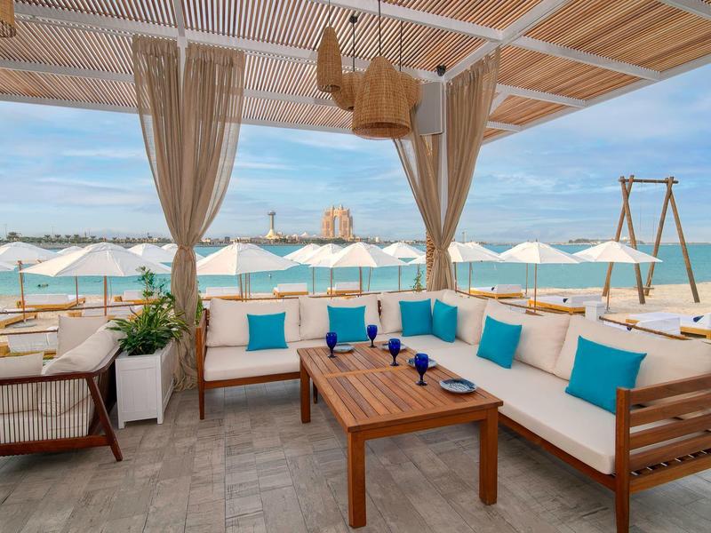 Offene Strandlounge mit Holzmöbeln, blauen Kissen, Sonnenschirmen und Blick aufs Meer.