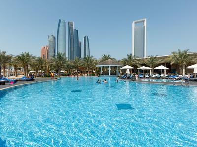 Großer Außenpool mit Sonnenliegen und Wolkenkratzern im Hintergrund bei sonnigem Himmel.