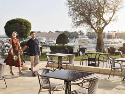 Paar läuft Hand in Hand auf einer Terrasse mit Tischen und Stühlen nahe einem Yachthafen.