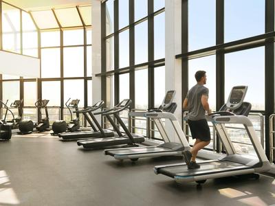 Moderner Fitnessraum mit großen Fenstern und mehreren Laufbändern, eine Person läuft auf einem davon.